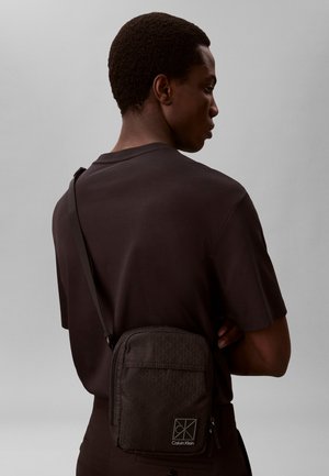 Bărbat văzut din profil, purtând un tricou negru și o geantă neagră de umăr cu logo-ul Calvin Klein, pe un fundal gri simplu.