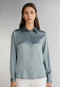 Blusa de satén en verde claro con un frente abotonado, mangas largas y un cuello clásico. Presenta una textura suave y un ajuste entallado.