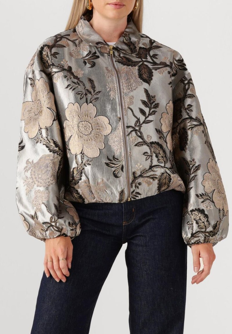Opgeblazen zilveren bomberjack met bloemenborduursel in beige en zwart. Ritssluiting en opstaande kraag, gecombineerd met donkerblauwe jeans.
