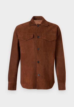 Man met kort blond haar die een bruine jas met knopen draagt, een beige overhemd, taupekleurige broek en tweekleurige bruine en beige loafers, staand.