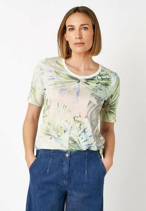 Frau mit kurzen blonden Haaren, die ein pastellfarbenes tropisches T-Shirt und blaue hoch taillierte Hosen trägt, steht mit den Händen in den Taschen.