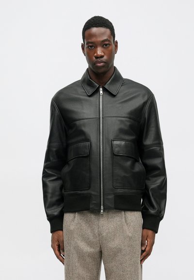 Giubbotto bomber in pelle nera con zip frontale, due tasche anteriori, polsini a costine e colletto classico. Texture liscia e raffinata.