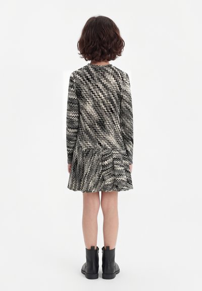 Bambino con capelli corti e ricci che indossa un vestito a maniche lunghe con motivo a zigzag bianco e nero e stivaletti neri alla caviglia, visto di spalle.