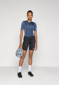 Maillot de cyclisme bleu marine à manches courtes, avec fermeture éclair à l'avant et logo ; cuissard noir rembourré ; casque gris tenu dans une main ; chaussures de cyclisme noires.