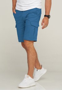Blaue Cargo-Shorts aus einem glatten Stoff, mit seitlichen Taschen und einer lockeren Passform, kombiniert mit weißen Sportschuhen.