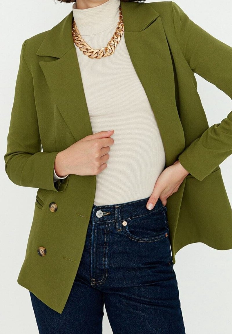 Oliwkowa dwurzędowa marynarka z szerokimi klapami i dwoma guzikami z przodu, noszona na kremowym prążkowanym golfie i ciemnych dżinsach.