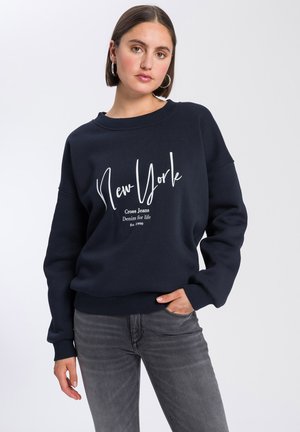 Granatowy sweter z białym napisem "Nowy Jork" i logo, z prążkowanymi mankietami i dołem, w luźnym kroju. Połączony z szarymi dżinsami.