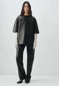 Donna che indossa un top e pantaloni in pelle nera oversize, guanti bianchi lunghi e tacchi aperti neri, in piedi contro uno sfondo chiaro e uniforme.