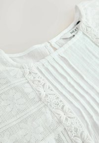 Blouse blanche avec des rayures texturées et des détails en dentelle délicate sur le devant. Le dos présente une petite ouverture en forme de clé avec une fermeture à bouton.