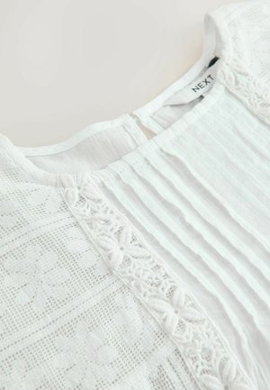 Vit blus med texturerade ränder och intrikata spetsdetaljer fram. Baksidan har en liten nyckelhålsöppning med knappstängning.