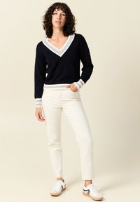Pull en V bleu marine avec col et poignets rayés gris et blanc, associé à un pantalon blanc et des baskets décontractées. Tissu lisse et coupe ajustée.