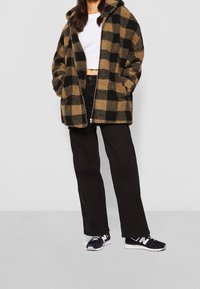 Rutet brun og sort fleecejakke med hætte, lynlås og sidetasker, parret med sorte bukser og sneakers.