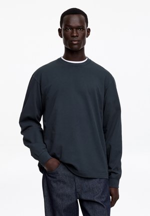 Mann trägt dunkles Langarmshirt und Jeans, steht mit einer Hand in der Tasche vor einem einfarbigen weißen Hintergrund.