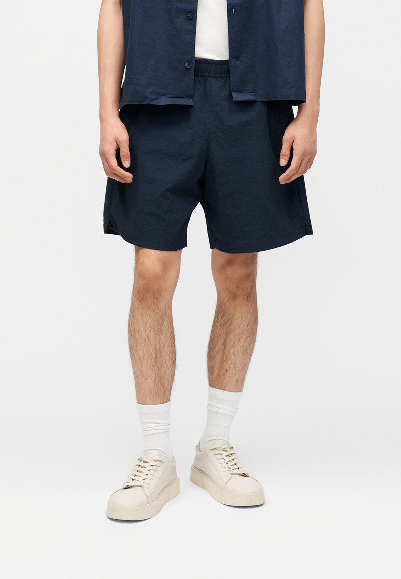 Άνδρας που φοράει σκούρα μπλε σορτς, λευκές κάλτσες και μπεζ αθλητικά παπούτσια, στέκεται μπροστά σε ένα λευκό απλό φόντο.