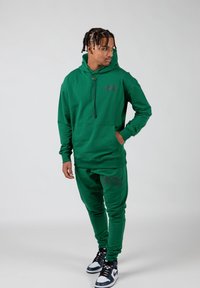 Groene katoen hoodie met een voorzak, trekkoord en logo. Gecombineerd met bijpassende joggingbroeken met logo, gedragen met zwart-witte sneakers.