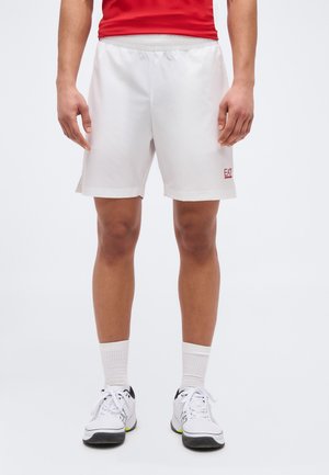 Mann trägt weiße EA7-Shorts, weiße Socken und weiße Turnschuhe, steht vor einem schlichten hellen Hintergrund.