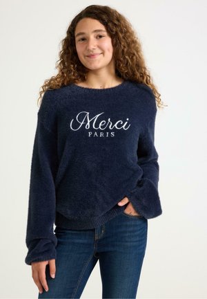 Granatowy puszysty sweter o okrągłym dekolcie i długich rękawach, z białym haftowanym napisem "Merci Paris" na przodzie. W zestawie z niebieskimi dżinsami.