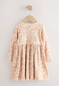 Robe à manches longues pour tout-petit avec imprimé floral rose et beige, suspendue à un cintre en bois contre un mur blanc.