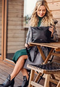 Černá kožená kabelka s dlouhým popruhem, obdélníkového tvaru, hladké textury, se malým logem. Osoba s kudrnatými vlasy ji drží, sedí na dřevěné židli.