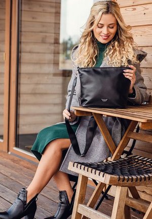 Černá kožená kabelka s dlouhým popruhem, obdélníkového tvaru, hladké textury, se malým logem. Osoba s kudrnatými vlasy ji drží, sedí na dřevěné židli.