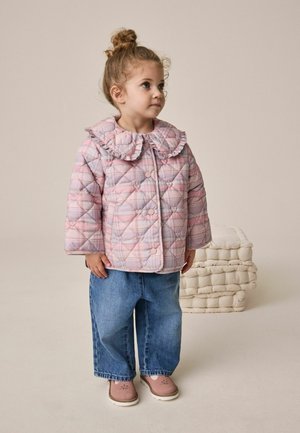 Lille barn stående iført en pink ternet quiltet jakke med flæsekrave, brede blå jeans og pink sko på en ensfarvet baggrund.
