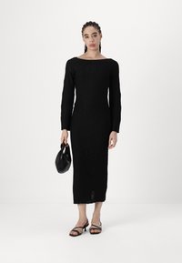 Vestido preto de manga longa com um padrão geométrico texturizado, até ao joelho, complementado por uma bolsa preta e sandálias pretas com tiras.