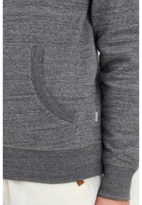 El sudadera gris de cuello redondo cuenta con un bolsillo curvado, puños acanalados y dobladillo, y una pequeña etiqueta de la marca. Fabricada en un tejido suave y texturizado.