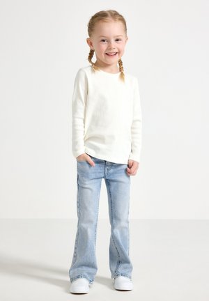 Vit långärmad ribbad skjorta som matchas med ljusblå utsvängda jeans och vita sneakers, stående mot en ljus bakgrund.