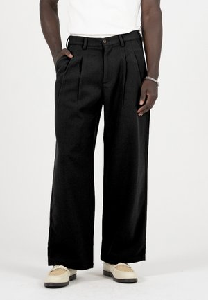 Man die losse zwarte geplooide broek, witte loafers en een wit shirt draagt, staand met één hand in de zak tegen een witte achtergrond.