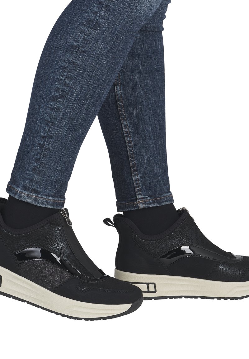 Personne portant un jean skinny foncé et des baskets slip-on noires avec des semelles épaisses blanches, debout sur un fond blanc.