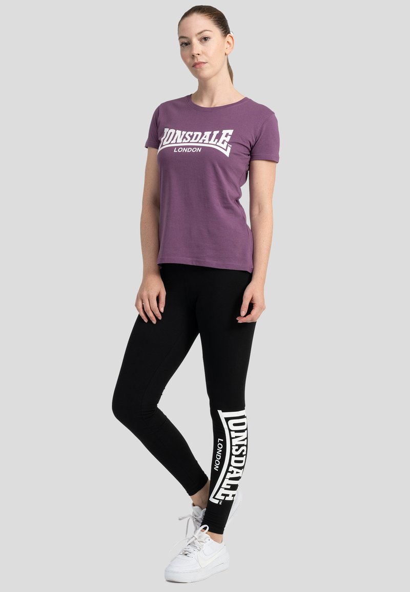 Joven de pie, vestida con una camiseta morada con el texto "Lonsdale London", leggings negros con texto blanco "Lonsdale London" y zapatillas blancas.