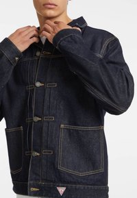 Dark denim jacket with a collar, front buttons, and two large pockets. Features contrasting yellow stitching and a logo patch at the hem.