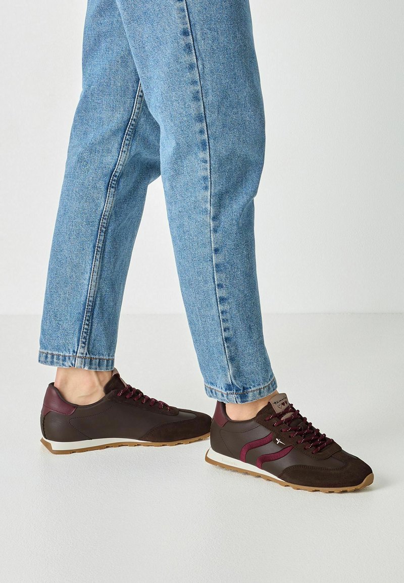 Personne portant des baskets marron et bordeaux avec des lacets violets et un jean en denim bleu clair, debout sur un sol clair.