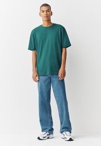 Žalia medvilninė marškinėliai laisvo kirpimo, trumpomis rankovėmis ir apvalia iškirpte, derinami su šviesiai plaunamais tiesaus kirpimo džinsais ir baltais sportbačiais.