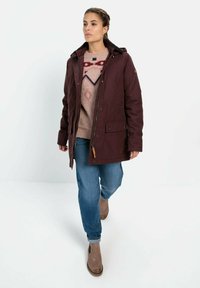 Burgunderfarbene wasserdichte Jacke mit Kapuze, Reißverschluss vorne und zwei vorderen Taschen, getragen über einem gemusterten Pullover und blauen Jeans. Braune Stiefeletten.