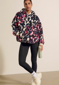 Fluffy, pullover hoodie in een levendige, veelkleurige luipaardprint; gecombineerd met donkere legging en chunky witte sneakers. Grijze drinkfles in de hand.