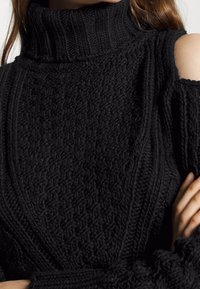 Femme portant un pull en tricot noir à col roulé avec une épaule découpée, mettant en valeur un motif torsadé texturé détaillé.