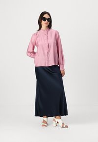 Rosa, volangert skjorte med knapper kombinert med et langt, gl glatt marineblått satin skjørt. Hvite hælsandaler fullfører antrekket. Mørke solbriller brukes.