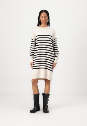 Robe pull rayée en crème avec des rayures marines. Présente un col rond et des manches longues. Portée avec des bottes noires jusqu'aux genoux.