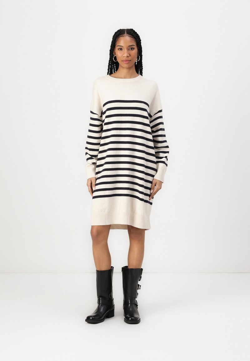 Striped knit sweater dress in cream with navy stripes. Features a round neckline and long sleeves. Worn with black knee-high boots.