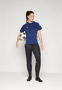 Jūras spēku sporta krekls ar baltām svītrām, melni pieguļoši bikses un melni futbola apavi. Tur rokās melnu un baltu futbola bumbu.