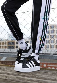 Musta-valkoiset Adidas Superstar -lenkkarit kumipohjalla, joissa on kolme raitaa, kultaiset yksityiskohdat ja teksturoitu varvaskuppi. Mustat jogger-housut, joissa on kukkakirjonta sivussa.