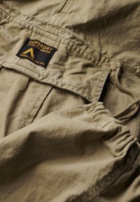 Khaki utility jack met een gestructureerde stof, trekkoordverstellingen en een opvallend zwart-gele label met het woord "Superdry."