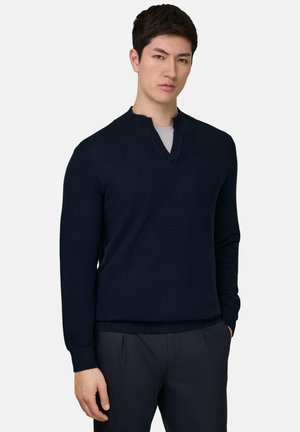 Mies, joka pukeutuneena tummansiniseen v-kaulaiseen neulepaitaan vaaleanharmaan paidan päällä ja tummansinisiin housuihin, seisoo yksivärisen taustan edessä.