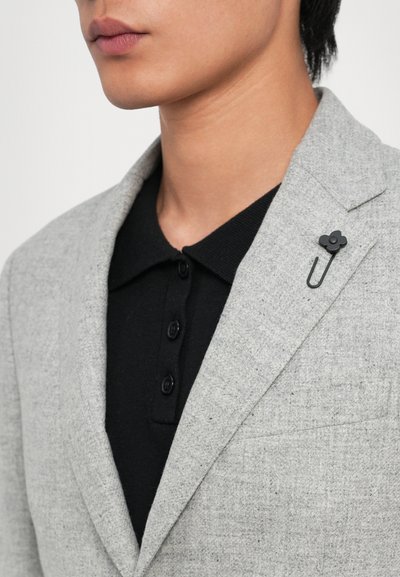 Light gray blazer with textured fabric, notched lapels, and a black flower pin on the lapel, worn over a black collared shirt with buttons.