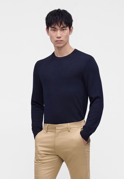 Mand med sort hår iført en mørkeblå, langærmet sweater og beige bukser, står med hænderne i lommerne mod en ensfarvet baggrund.