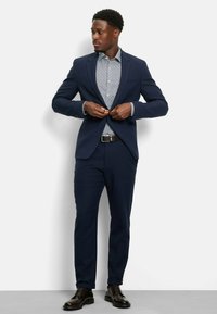 Marineblauer Anzug mit einem taillierten Blazer und Hose, kombiniert mit einem gemusterten hellblauen Hemd. Dunkle Lederschuhe runden den Look ab, während die Hände den Blazer anpassen.