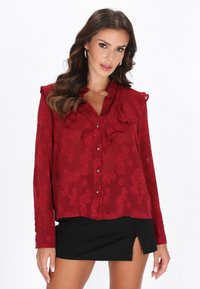 Blusa roja con estampado, cuello con volantes, mangas largas y cierre de botones. Tela semitransparente con textura floral, combinada con una falda negra.