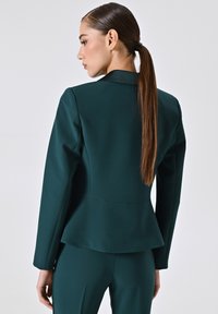 Blazer peplum in tessuto verde intenso, con spalle sartoriali, corpetto aderente e una texture liscia. La parte posteriore presenta una silhouette pulita.
