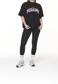 Černé oversized tričko s bílým nápisem "PEGADOR", v kombinaci s černými legínami. Bílá sportovní ponožky a šedé sportovní boty dotvářejí vzhled.